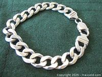 Chunky thick sterling silver men's bracelet laid in a circular formation on dark green cloth, showing thick, flat curb chain links and lobster clasp.