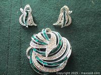Front view of brooch and pair of matching snap-on earrings showing clear and emerald green rhinestones in swirled design on rhodium plated metal.