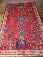 Full view of red wool rug laid on hardwood floor showing overall field, border and fringe