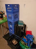 Stack of 4 blue Scotsburn milk crates, 2 black crates, green lidded bin, smaller containers, and shopping bags piled near a black cart and table.