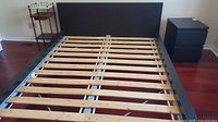 Black wooden Ikea bed frame with wooden slats and headboard in room with reddish hardwood floor, showing full frame and slats arrangement.