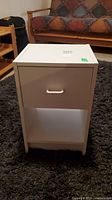 Front view of small white end table showing drawer and open shelf on dark carpet