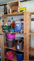 Four wooden shelves with various plastic buckets, a blue metal watering can, Scotts spreader, garden hose nozzle, small plastic pots, gardening gloves, and a wooden crate on top shelf.