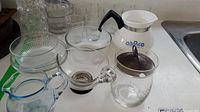 Photo showing various clear glass bowls, a glass vase, nine small water glasses, and a CorningWare teapot with black handle and blue floral design, missing lid, along with creamers and sugar dish.