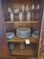 Photo shows upper shelf with wine glasses, cocktail glasses, thin lipped juice glasses. Middle shelf contains stacked white plates, some smaller plates, and Arabia serving plate. Bottom shelf shows two covered glass bowls with lids.