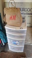 Photo of a white plastic drawer organizer with four translucent drawers stacked vertically, a brown paper bag on top with a red H&M logo visible.