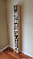 Front view of tall wooden CD media tower filled with CDs, standing against a beige wall with hardwood floor.