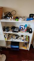 Wide view of shelf showing mixture of office supplies and electronic items including alarm clocks, adapters, tape dispenser, and boxes.