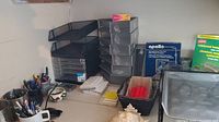 Wide view of office supplies on countertop showing multiple black plastic drawer organizers, pens in containers, loose paper, Apollo photocopy paper box, and other items