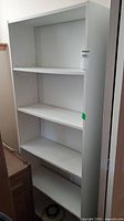 Front view of white shelf unit showing three shelves and adjustable shelf peg holes on the sides.