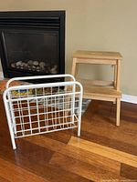 Wooden two step stool and white metal magazine rack next to fireplace on wooden floor