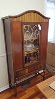 Front view of vintage wooden china cabinet showing curved top, glass door with decorative wooden grille, and lower drawer.