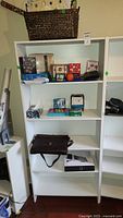 White shelving unit with multiple shelves containing assorted office supplies, copy paper, gift bags, and a basket on top.