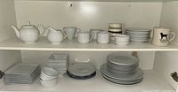 Shelf full of white round plates stacked along with square white bowls and plates, small white dishes and blue plastic plates on the left. Left to right visible items include larger white plates, smaller white plates, and various square and round bowls.