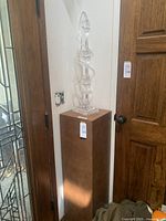 Full view of acrylic sculpture mounted on brown base placed in corner near door.