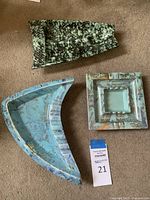 Three ashtrays shown on carpet floor, light blue and gold boomerang shape, black and green speckled boomerang, and square marbled ashtray.