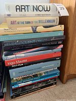 Stack of 17 assorted art and architecture books on floor next to wooden furniture.