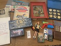 Overview of lot layout showing Barbie doll, board games, Magic The Gathering cards, framed botanical art, specimen display, coin collection, and books