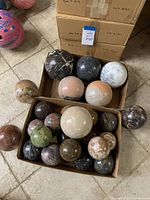 Multiple polished spheres made from marble and other stones in two cardboard boxes and on the ground, showing a variety of earth tone colors and veining.