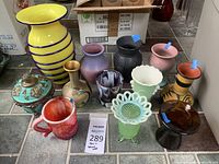 Lot of 12 assorted decorative pottery and flower bases viewed from front on tiled floor, showing variety in colors and styles with tallest yellow vase and smallest red cup.