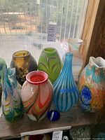 Seven decorative glass vases, colorful and patterned, displayed on wooden shelf near window.