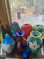 Several decorative glass flower vases of varied colors, shapes and sizes with a blue diamond-shaped paperweight in front.