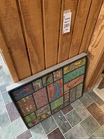 Full view of framed art resting against a wood-paneled wall on tile floor, showing the artwork and frame.