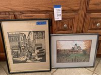 Two framed art prints placed side by side on wooden floor against wood cabinet. Left print is black and white urban scene, right print is a color image of house with green lawn.