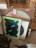 Group of framed artworks leaning against wall, including green-blue geometric piece, pastel circle, op art circle, and assorted prints