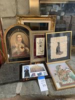 Wide shot showing all framed art pieces grouped together on floor against wall; includes religious portrait, Egyptian cat papyrus, traditional cultural artworks, and photographic prints.