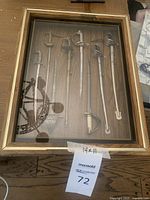 Full view of the wooden shadowbox frame containing seven miniature swords displayed vertically with metal blades and varied handles.