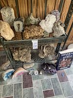 Two-tier metal shelf displaying multiple mineral specimens including rough crystals, geode halves, a white botryoidal cluster, a brown tan desert rose cluster, agate slices, small quartz and amethyst pieces, and a "Minerals of the World" book.