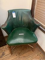 Front view of the green leather chair showing curved backrest, padded seat, brass nailhead trim, and wooden legs on wood floor.