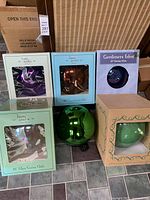 Photo showing six glass gazing globes in their original boxes arranged on a tiled floor. Visible colors include green, purple, bronze/copper tone, and light green iridescent. Most boxes labeled with 'home garden décor' branding and one with 'Gardeners Eden.'