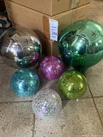 Photo showing six garden gazing globes including two large mirrored and four smaller crackled glass globes in various colors placed on the floor near cardboard boxes.