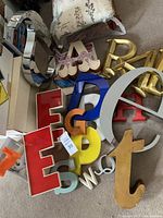 Collection of various decorative letters of multiple sizes and colors, made from wood, metal, and acrylic, arranged on floor with visible diverse shapes and finishes.