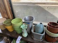 Various ceramic planters, pots, and vases of different sizes and colors shown on a wooden shelf against a window.