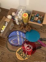 Wide view showing all items including multiple boxes of balls, wooden puzzles, paddle ball sets, and tennis rackets with cover.