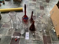 Group of scientific glassware including clear and amber volumetric flasks, beakers, graduated cylinders, and thermometers arranged on floor tiles.