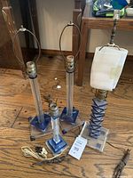 Four table lamps on wood floor, three with clear acrylic cylindrical bodies and blue square bases, one blue geometric pattern lamp, cords visible, metal bulb holders at top, no lampshades.