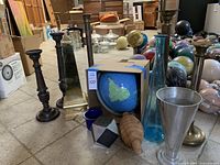 Photo of multiple tall candlestick holders, a globe, glass bottles, and other decorative objects on floor with boxes and other items in background.