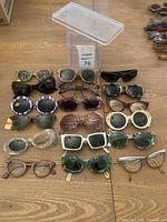 Full lot of vintage ladies glasses and sunglasses arranged in 5 rows on a wooden surface with plastic container in background.
