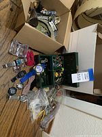 Overall view showing watches, green box of accessories and cardboard box of appliqués on wooden floor