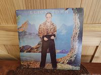 Front cover of Elton John Caribou LP showing Elton John standing outdoors in colorful background with slight corner and edge wear.