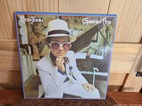 Front cover of Elton John Greatest Hits album showing Elton John in white suit and sunglasses beside a piano.