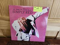 Front cover of Simply Red "A New Flame" 12 inch vinyl LP showing pink background with image of person in white with curly hair and a yellow vendor sticker on front.