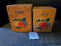 Photo showing two nesting wooden canisters labeled 'Flour' and 'Sugar'. Each has a painted fruit design and wooden lids with round handles on top.