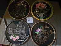Four vintage metal state souvenir trays arranged on a cloth surface, showing Texas, Tennessee, West Virginia, and New York models with detailed maps and floral decorations.