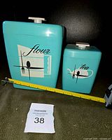 Two turquoise hard plastic canisters labeled flour and tea with mid-century modern graphic designs and white lids, placed side by side with measuring tape showing size comparison.