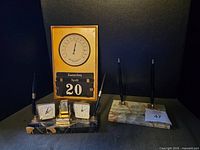 Marble barometer and clock, desk calendar showing April 20, with date and month spinning functions visible, set on black surface with pens.